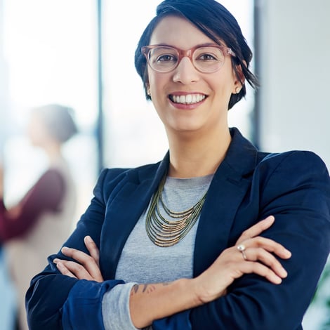 Smiling professional woman with glasses stands with arms crossed in an office setting.
