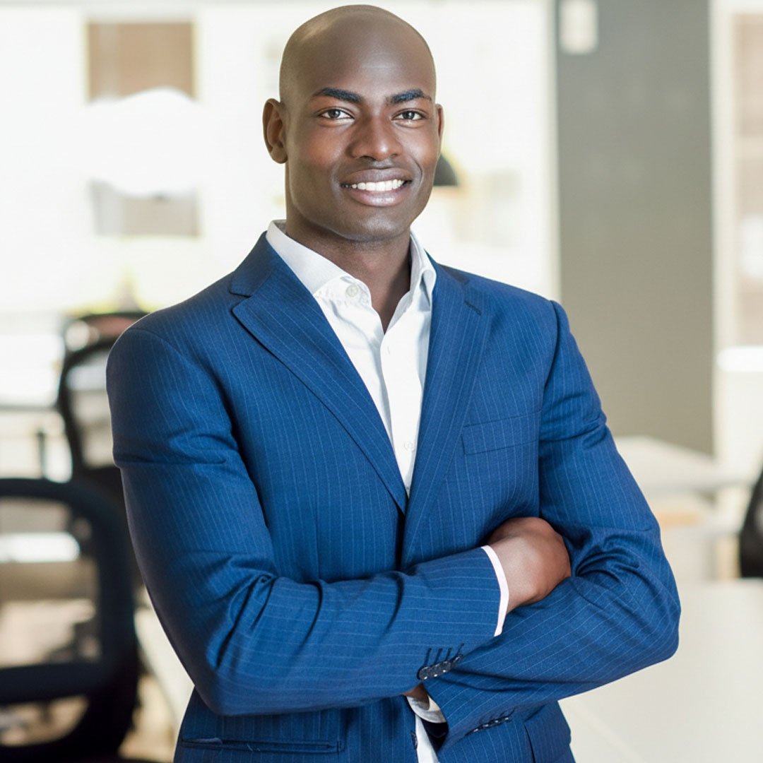 Smiling businessman in a suit stands with arms crossed in an office.