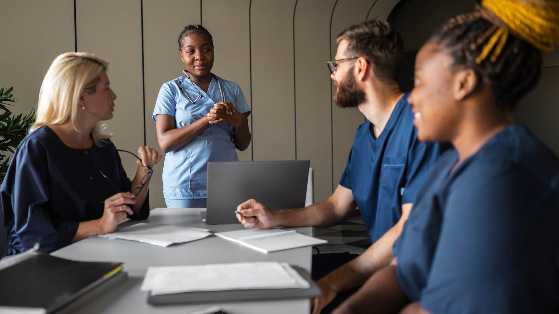 Healthcare professional presenting to a team of colleagues in scrubs around a table with a laptop.